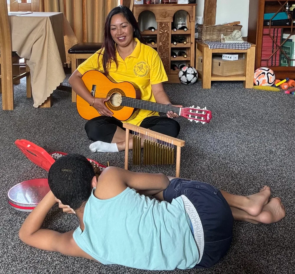 Tess playing guitar during a home music therapy session, with chime bars close by as a new client settles in